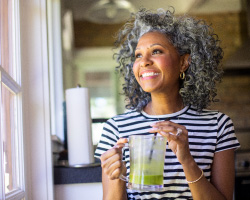 A smiling woman holds a smoothie while looking out a window.