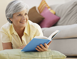 A woman in a yellow shirt with silver hair looks up from her book and smiles.