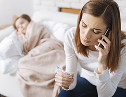 A woman looks at a thermometer in concern and holds a phone to her ear. Behind her, a young girl looking ill is wrapped in blankets. 