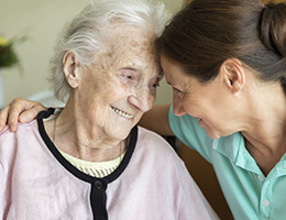 A younger woman has her arm around the shoulders of an older woman. The two women are smiling at each other as their foreheads touch.