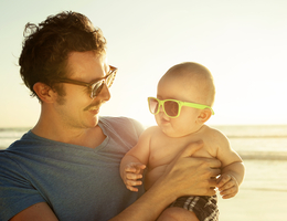  A man holds a baby on a beach. Each wears sunglasses.