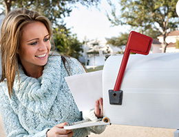 A woman in a blue sweater is standing outside and pulling mail out of a white mailbox.