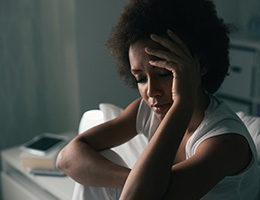 A woman sits in a dark bedroom with her eyes closed.