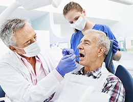 A dentist looks in a man's mouth with a dental hygienist in the background. 