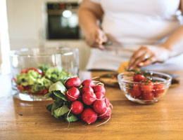 A wooden countertop with a bundle of radishes, a bowl of tomatoes and a half-filled salad bowl. In the background, a woman chops salad ingredients.
