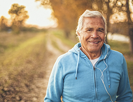A man with white hair, wearing a turquoise hoodie and white earbuds, walks along an outdoor trail at sunset.