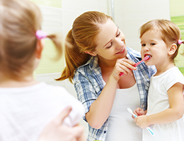 A woman in a blue plaid shirt helps her young daughter brush her teeth with a pink toothbrush. 