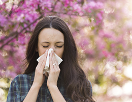 A woman blows her nose while standing next to a tree with pink flower blossoms on it.
