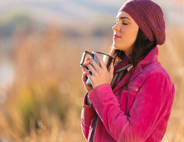 A woman in a hat, scarf and coat holds a mug in both hands.