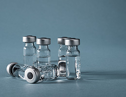 Several glass vials of clear vaccine liquid sit on a blue background. 