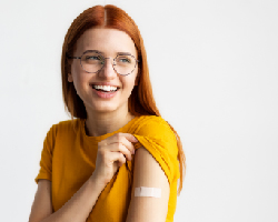 A smiling adolescent girl lifts the sleeve of her T-shirt to display an adhesive bandage.