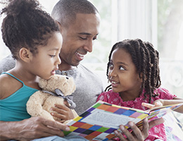 A father holds his two young daughters on his lap, reading to them from a brightly colored book. One girl points at a page, the other cuddles a teddy bear.