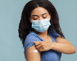 A woman in a mask raises her short sleeve to show an adhesive bandage.