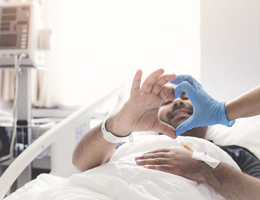 A man in a hospital bed reaches up, his hands meeting gloved hands to form a heart shape