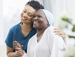 A woman in teal scrubs puts her arm around a woman in a white headscarf. They tilt their heads together and smile. 